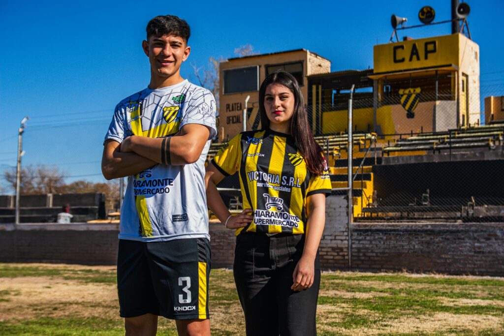 Two young adults in soccer jerseys pose confidently in front of a stadium on a sunny day.